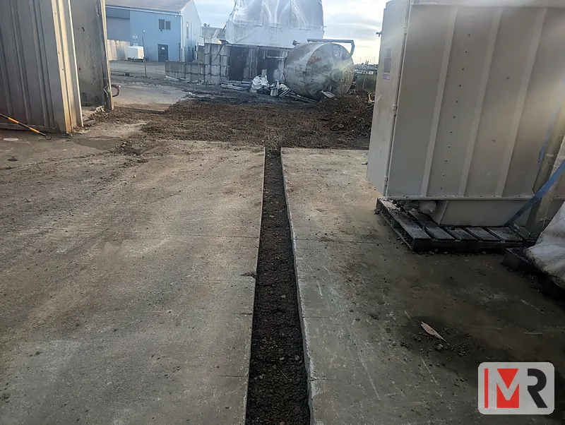 Cable trench cut through a concrete yard beside equipment